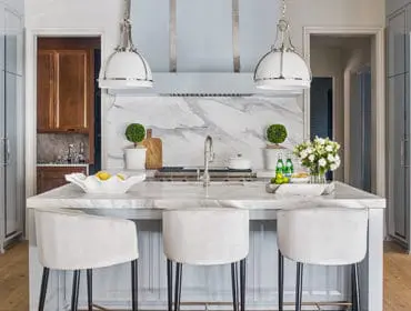 Light blue kitchen with marble countertops and backsplash, white pendant lights, and three white velvet bar stools at the island.
