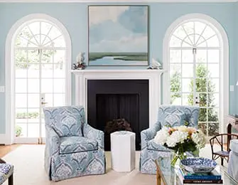 Living room with light blue walls, fireplace with black surround, two upholstered armchairs in blue and white damask pattern, and arched windows with white mullions.