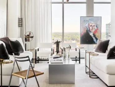 Modern living room with white sofas, a silver coffee table, and a portrait of George Washington above a large window with a city view.