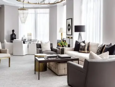 Living room with neutral-toned furniture, including a cream sofa with decorative pillows, gray armchairs, and a cream accent chair with gold legs. A dark wood coffee table sits in front of the sofa.
