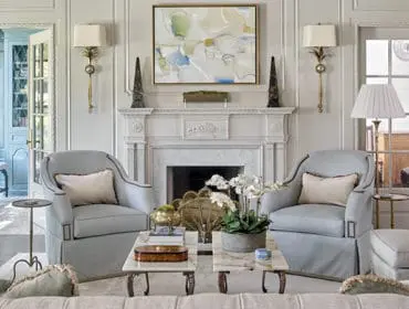 Light blue upholstered armchairs flank a white marble fireplace with abstract art above. A sunroom with a bench is visible through French doors on the left.