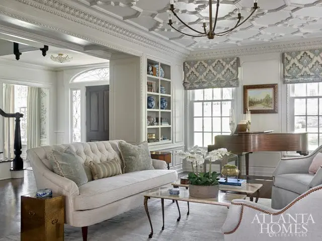 Living room with tufted sofa, grand piano, ornate ceiling, and built-in bookshelves filled with blue and white porcelain.