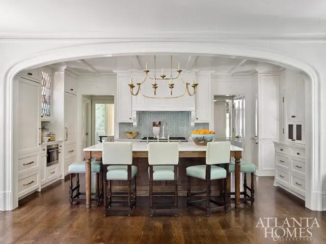 White kitchen with coffered ceiling, marble island with wooden base, light blue and white checkered bar stools, and a gold chandelier.