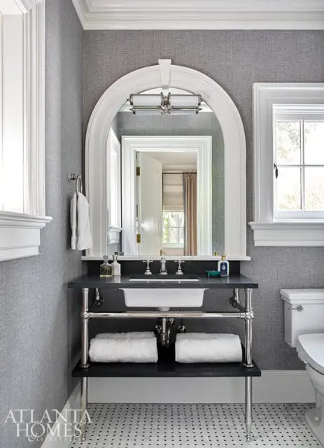Gray textured wallpaper bathroom with arched mirror over a dark stone vanity with white sink, chrome fixtures, and two white towels on the shelf below.