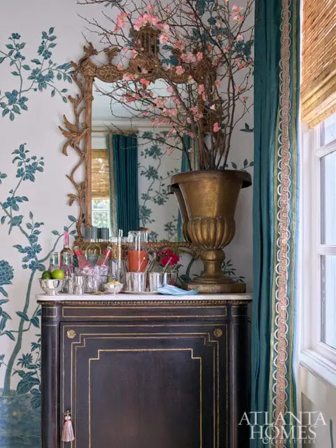 Dark wood bar cabinet with gold inlay topped with marble, set with bottles, glassware, and a vase of pink cherry blossoms. Blue floral wallpaper and teal curtains frame the scene.