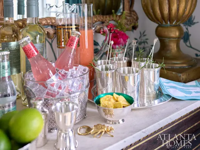 Silver jigger and bottle opener on a marble countertop next to a bowl of banana chips and silver cocktail glasses.