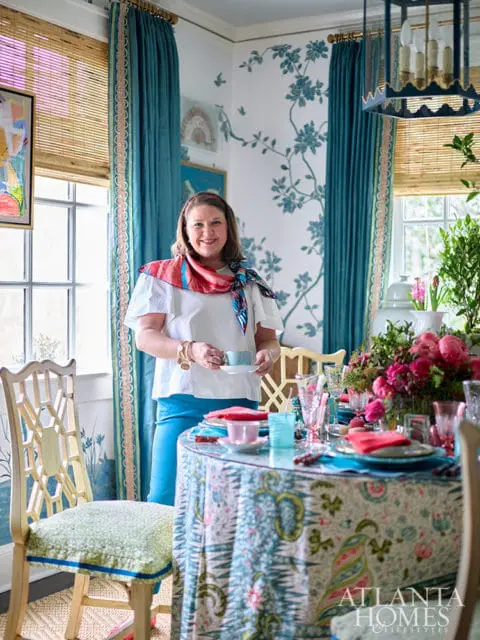 Woman in white shirt and blue pants holding a teacup in a dining room with blue floral wallpaper, teal curtains, and a patterned tablecloth.