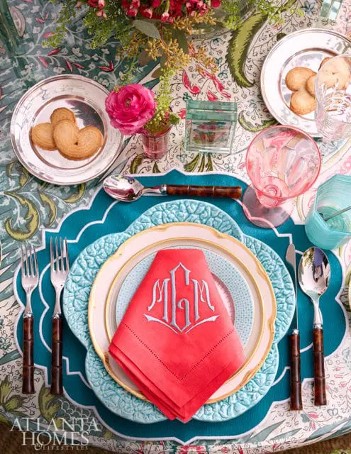Table setting with coral embroidered monogrammed napkins, blue textured chargers, gold rimmed plates, silver cutlery with bamboo handles, and heart-shaped cookies on silver chargers.