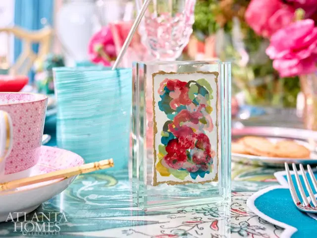 Close-up of a small framed abstract watercolor painting with pink, red, and blue floral shapes, set on a table with a pink teacup and a turquoise glass vase.