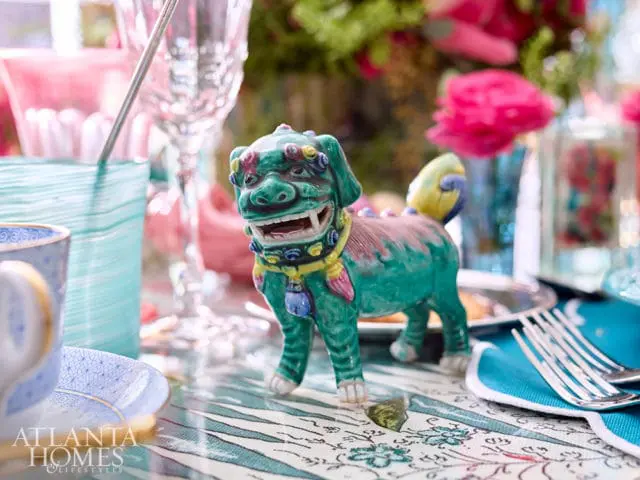 Green ceramic foo dog figurine with yellow and blue accents on a patterned tablecloth, surrounded by glassware and floral arrangements.