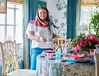 Mallory Mathis stands in a dining room holding a teacup and saucer. The room features a table set for tea with floral linens, pink glassware, and a centerpiece of pink peonies.
