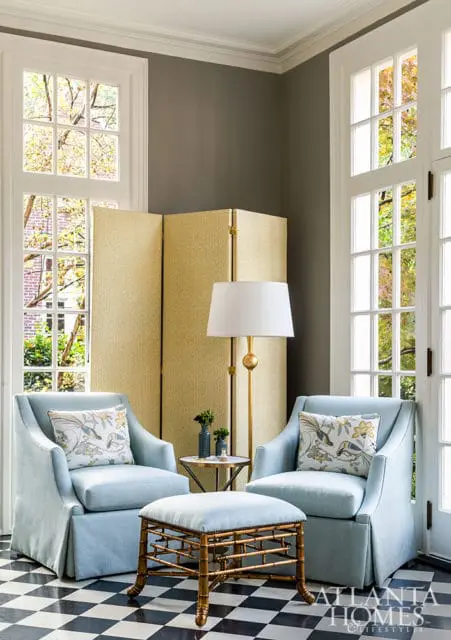 Light blue upholstered armchairs with decorative pillows flank a bamboo-style ottoman on a black and white checkered floor. A gold floor lamp stands behind a patterned folding screen.