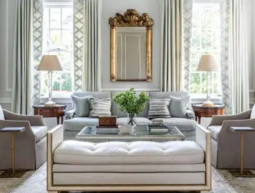 Light blue sofa with patterned pillows, flanked by upholstered armchairs and side tables. A gold ornate mirror hangs above the sofa.
