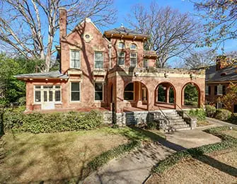 Victorian-style brick house with arched entryway, stone foundation, and large windows, surrounded by mature trees and manicured hedges.