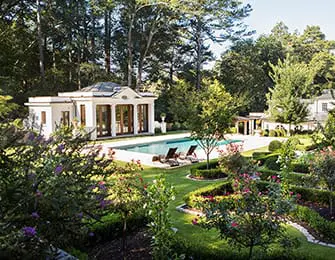 Neoclassical pool house with columns and a mansard roof overlooks a swimming pool and manicured gardens with lush trees and flowering bushes.