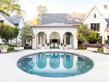 White stucco mansion with a grey shingle roof, featuring a kidney-shaped pool, arched loggia with outdoor seating, manicured hedges, and lush landscaping.