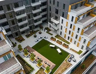 Modern apartment complex courtyard with artificial turf, seating areas, planters, and multi-story buildings with balconies.