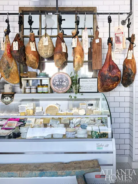 Deli display case filled with various cheeses and cured meats hanging above, white subway tile walls, and a rustic wooden bench in the foreground.