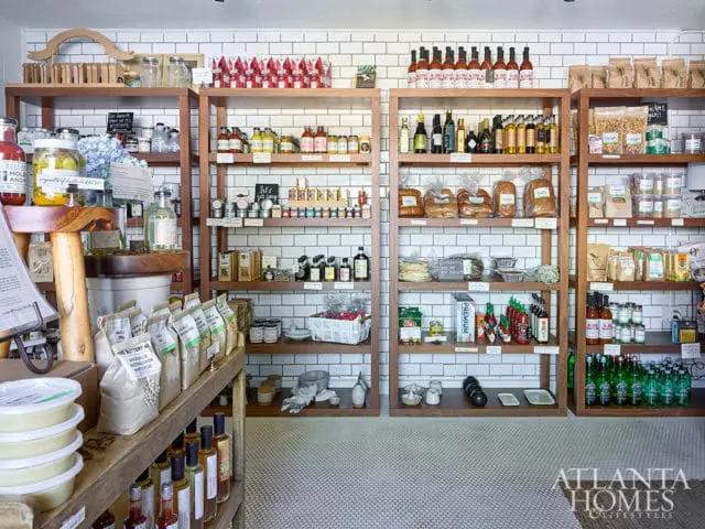 Wooden shelves filled with jars, bottles, bread, and packaged goods against a white subway tile backsplash.
