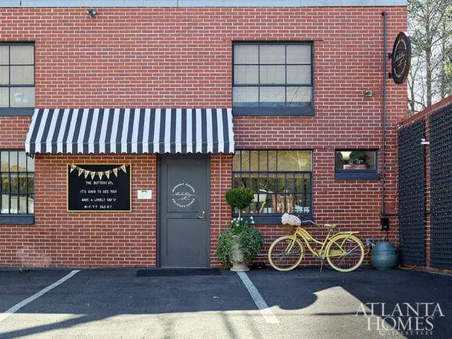 Red brick building exterior with a black and white striped awning over a door and window. A yellow bicycle with a basket of flowers is parked outside.