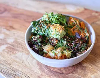 Bowl of food with beets, sweet potatoes, kimchi, avocado, and fresh herbs on a wooden cutting board