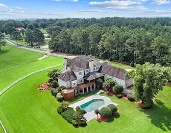 Large white house with gray roof, multiple dormers, and a screened-in porch, set on a large green lawn with a swimming pool and surrounding trees.