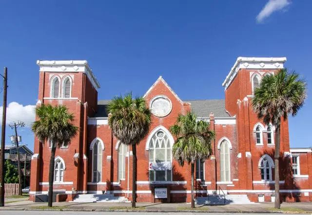 Red brick church with white trim, arched windows, and two towers, surrounded by palm trees under a bright blue sky.