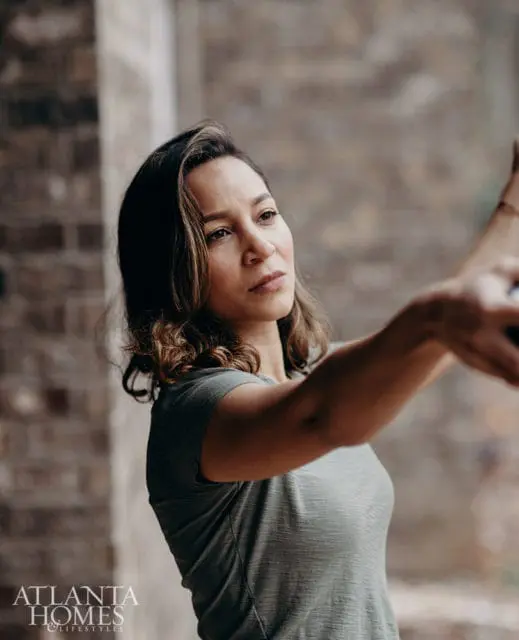 Woman with shoulder-length wavy brown hair wearing a green t-shirt, looking to the right with her arm extended.