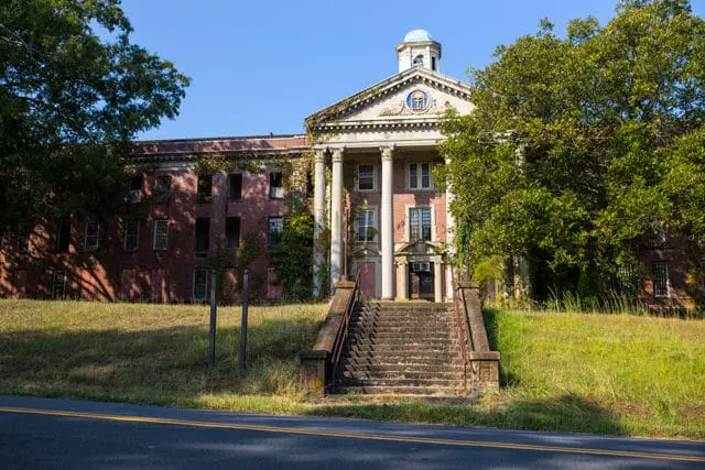 Abandoned brick building with a central portico, large columns, and a domed cupola, surrounded by overgrown grass and trees.