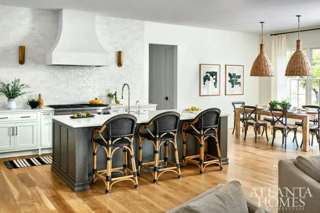 Modern kitchen with white subway tile backsplash, a large range hood, dark gray island with black rattan bar stools, and a dining area with a wooden table and chairs.