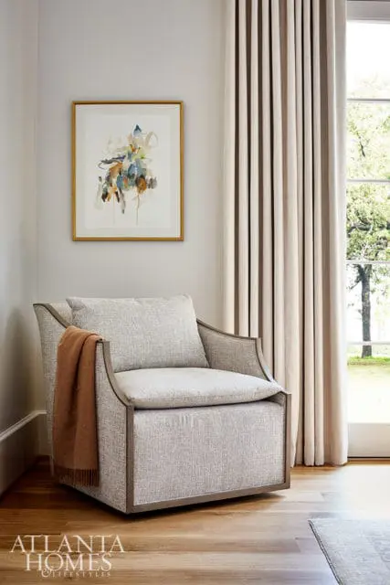 Gray upholstered armchair with a rust-colored throw blanket draped over the arm, next to a framed abstract painting and floor-to-ceiling curtains.