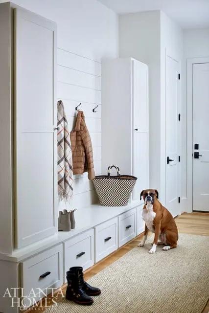 White mudroom with shiplap wall, built-in bench with drawers, tall cabinets, and a boxer dog sitting on a woven rug.
