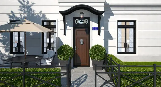 White house with a dark wood front door, black-paned windows, and a black arched awning with a lantern. A patio table with an umbrella and chairs sits to the left, with a black metal fence in the foreground.