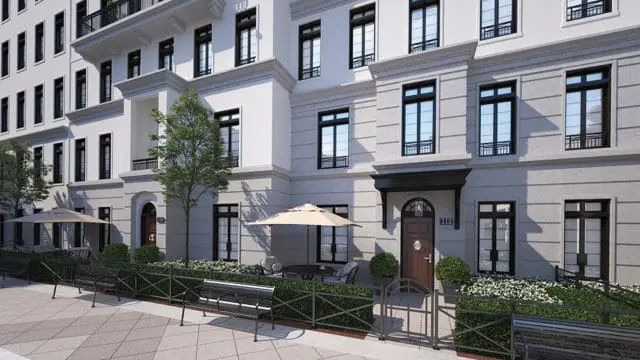 Row of townhouses with light gray stone facade, white trim, and black-framed windows. A paved walkway with benches leads to a small patio with an umbrella and seating.