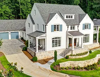 White modern farmhouse with stone accents, gray shingle roof, blue shutters, and a three-car garage. Lush landscaping surrounds the property.
