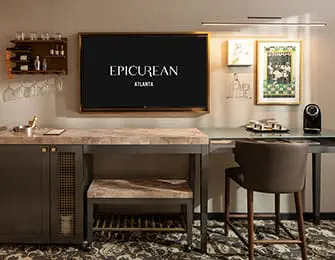 Hotel room with a large TV displaying 'EPICUREAN ATLANTA', a marble-topped island, a desk with a coffee maker, and a bar area with wine glasses.