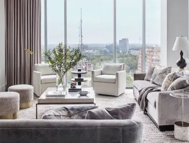 Living room with plush gray sofas, cream armchairs, and a marble-topped console table, overlooking a city skyline through floor-to-ceiling windows.