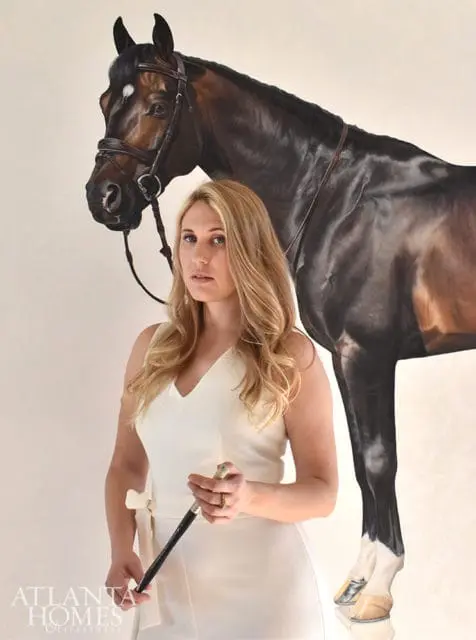 Woman in a white dress holding a riding crop, standing in front of a painting of a dark brown horse.