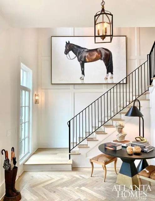 Foyer with herringbone wood floors, a large painting of a brown horse, a black metal staircase railing, and a round table with decorative objects and books.