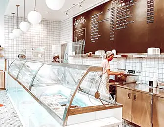 Clean, bright ice cream shop interior with white tile walls, a glass display case filled with ice cream, and a large chalkboard menu above the counter.