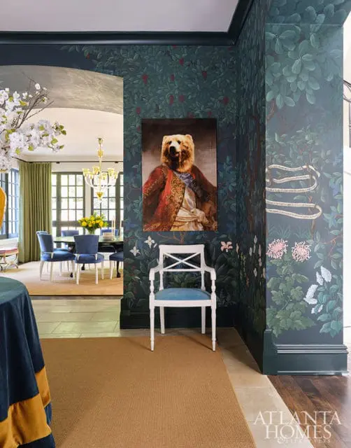 Hallway with dark floral wallpaper, a portrait of a bear in a red coat, and a white chair with blue upholstery. A dining room with blue chairs is visible through an archway.