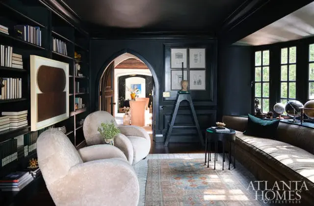 Dark moody library with floor-to-ceiling bookshelves, plush cream armchairs, a curved brown sofa, and an arched doorway leading to another room.