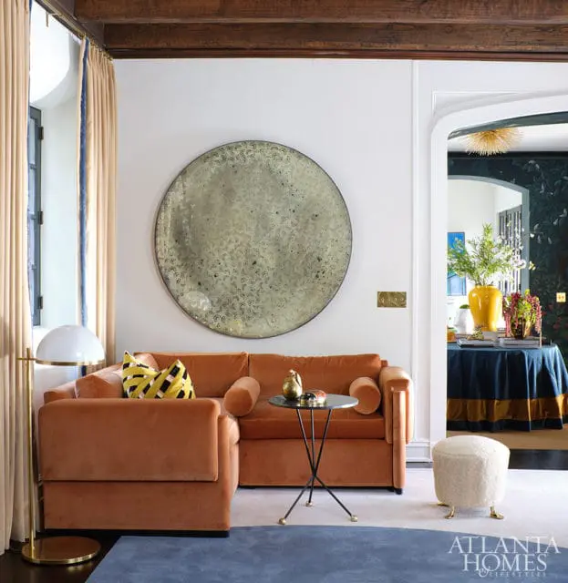 Living room with burnt orange velvet sectional sofa, round textured mirror, and a small black side table with a white boucle ottoman.