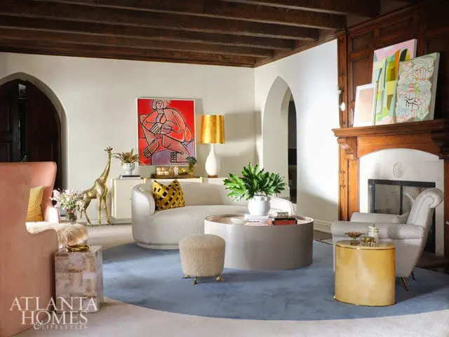 Living room with cream walls, dark wood ceiling beams, arched doorways, fireplace with wood mantel, and a large blue rug. Furnishings include a white bouclé sofa, pink armchair, patterned accent chair, and various decorative objects.