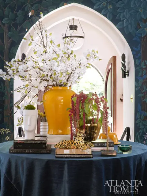 Table with a dark blue tablecloth displaying books, a leopard figurine, a white bust vase, a large yellow vase with white flowers, and a gold vase with red flowers. The background features an arched doorway and dark floral wallpaper.