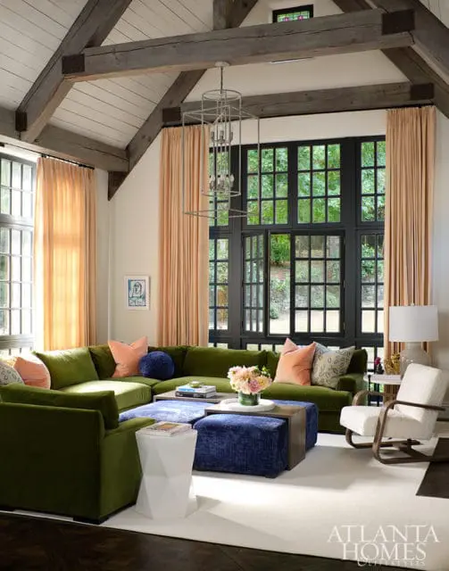 Living room with vaulted wood-beamed ceiling, large black-paned window overlooking greenery, and a large sectional sofa in olive green with blue and coral accents.
