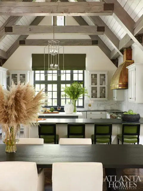 Vaulted ceiling with exposed wooden beams above a white kitchen with a brass range hood, dark countertops, and green bar stools. A large dining table with cream chairs is in the foreground.