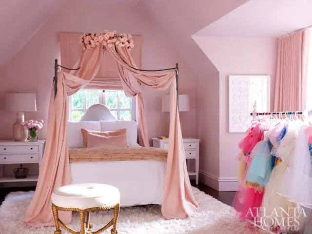 Pink bedroom with a canopy bed draped in sheer pink fabric, a white shag rug, and a rack of colorful children's dresses.