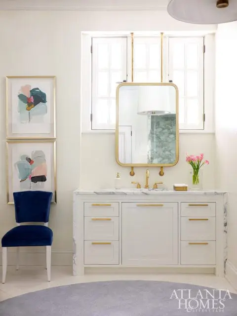White bathroom vanity with marble countertop and gold hardware, flanked by two framed abstract art pieces and a navy velvet chair.