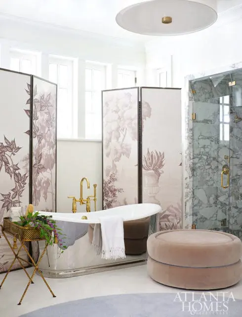 White bathroom with freestanding tub, brass fixtures, marble shower, decorative screens, and plush ottoman.